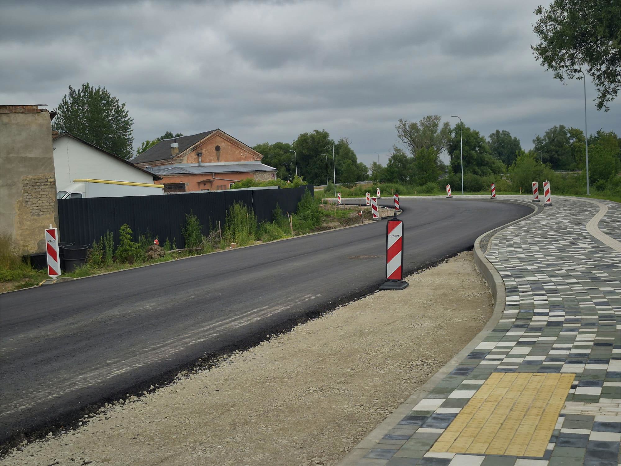 Jauns asfalta ceļš ar sarkani-baltām brīdinājuma barjerām un bruģētu ietvi, blakus vecai ēkai.
