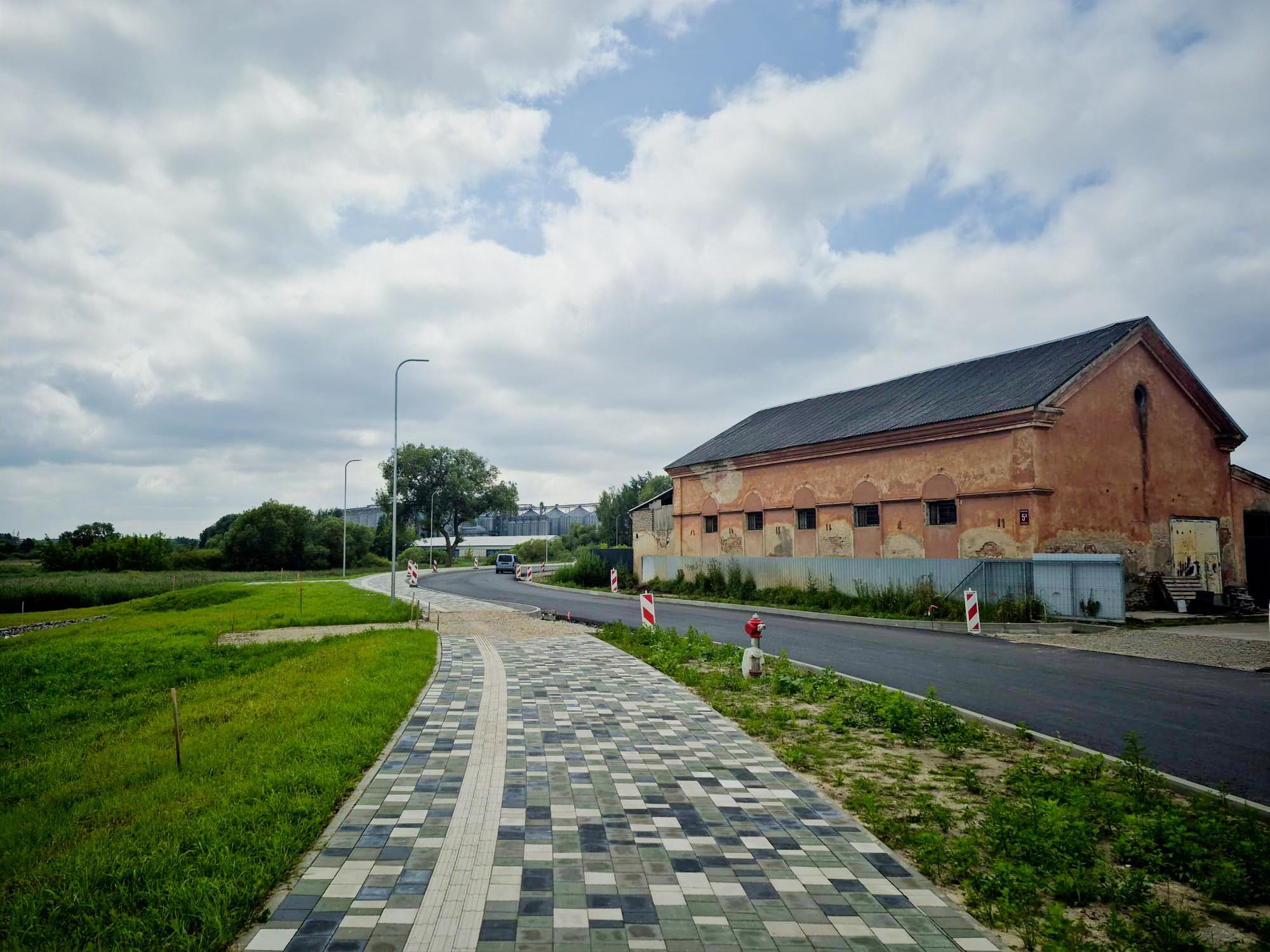 Gājeju celiņš ved uz vecu, sarkanīgu noliktavas ēku un jaunu asfalta ceļu ar barjerām.