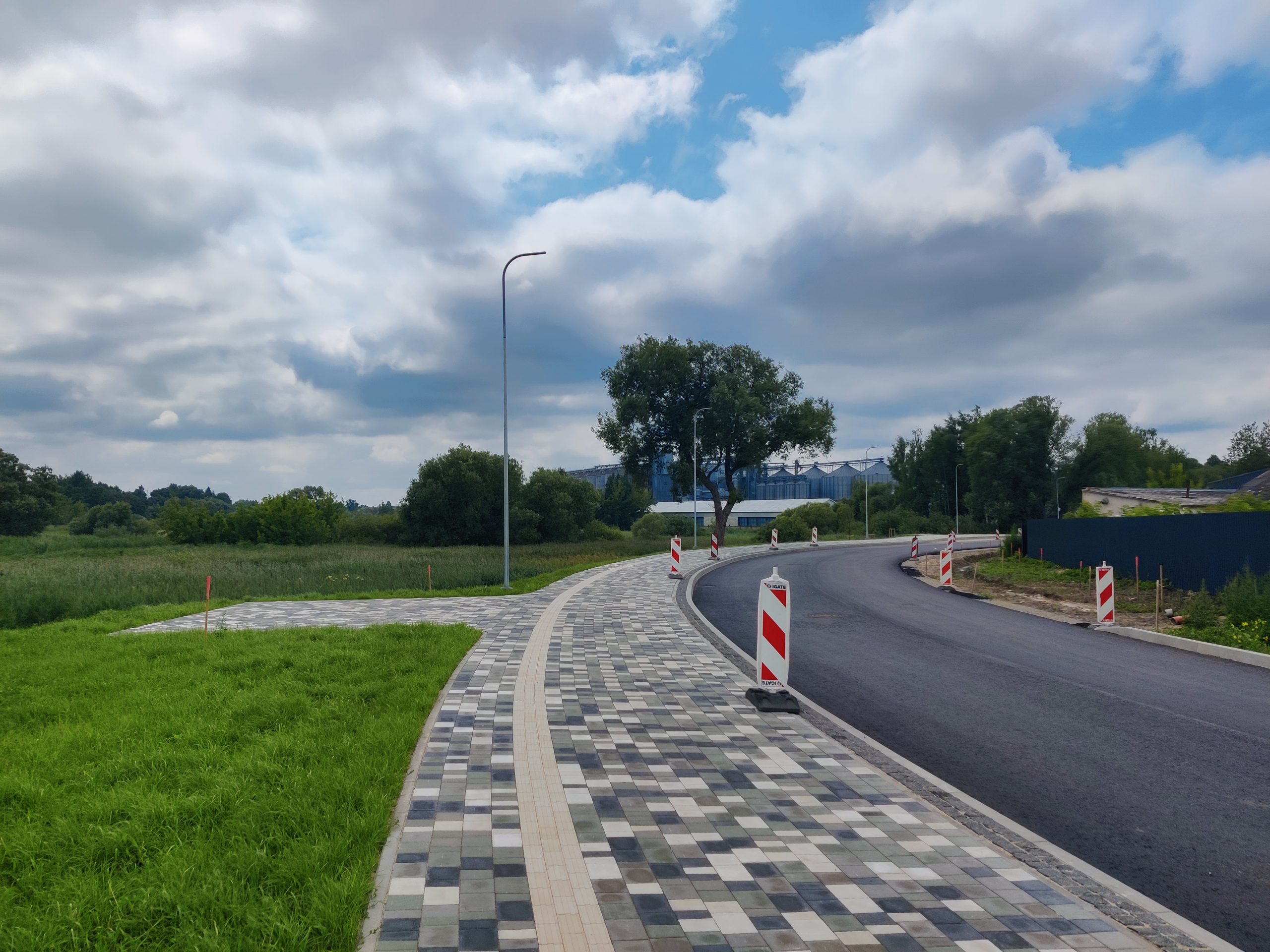 Bruģēta ietve un asfalta ceļš ar brīdinājuma barjerām, ved uz koku grupu un rūpniecisku fonu.