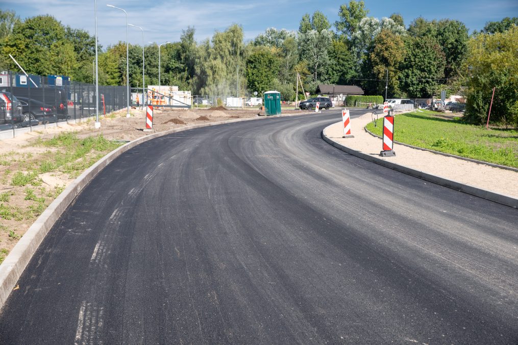 Jaunizbūvēts, gluds asfalta ceļa līkums ar apmalēm un ceļa darbu norobežojošajiem stabiņiem.