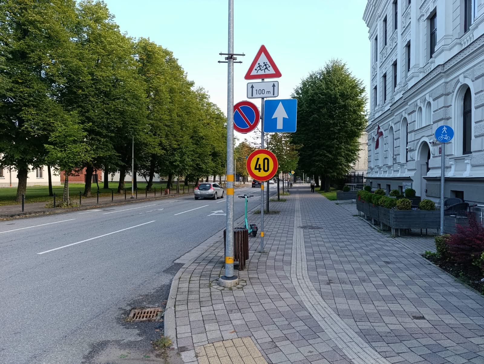 Pilsētas ielas skats ar ietvi un ceļa zīmju stabu, uz kura redzams dzeltenais ātruma ierobežojums 40 km/h, gājēju pārejas brīdinājums un citas ceļa zīmes.