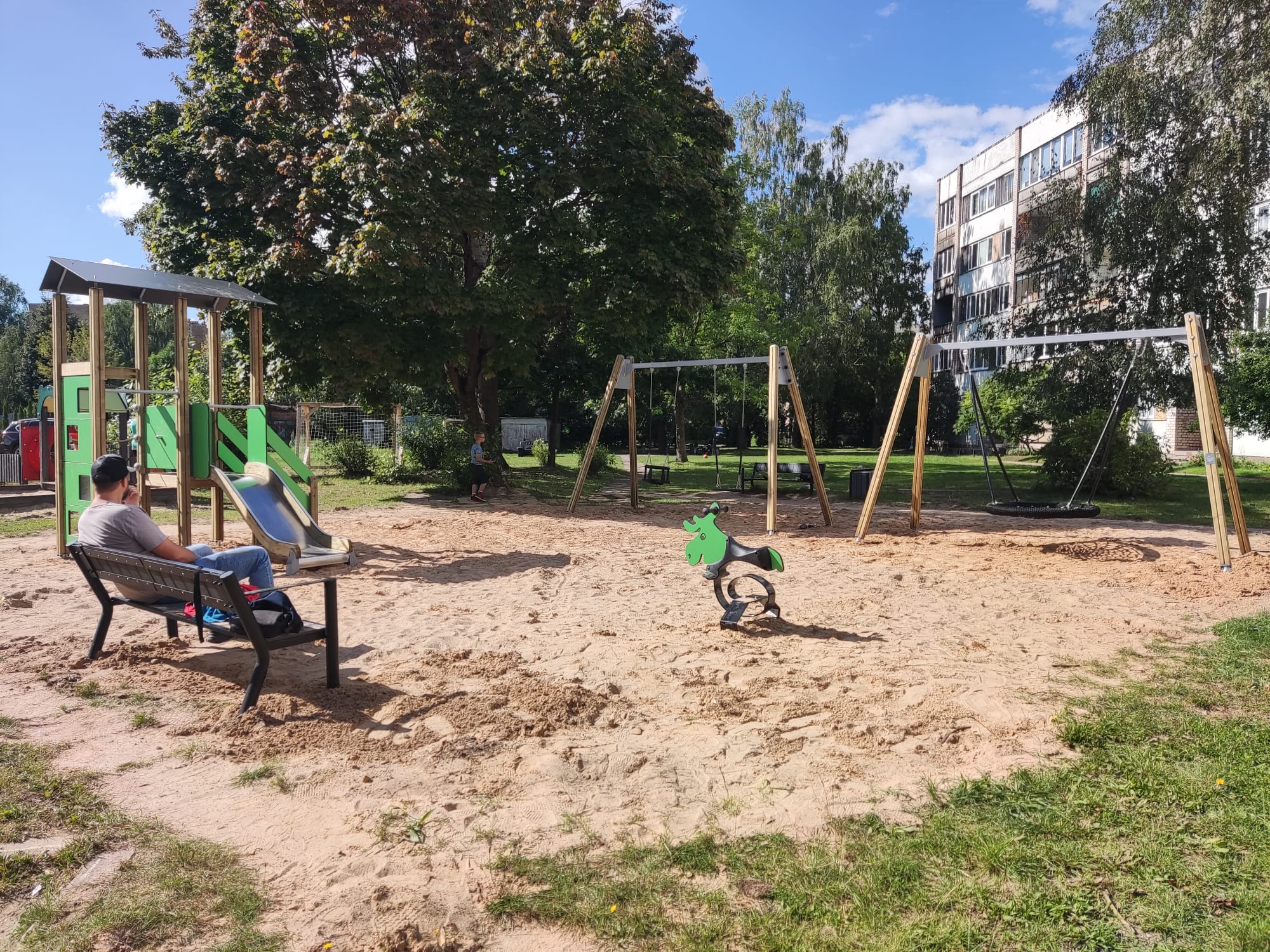 Bērnu laukums ar smiltīm, slidkalniņu, šūpolēm un pieaugušo uz soliņa, fonā daudzdzīvokļu māja.