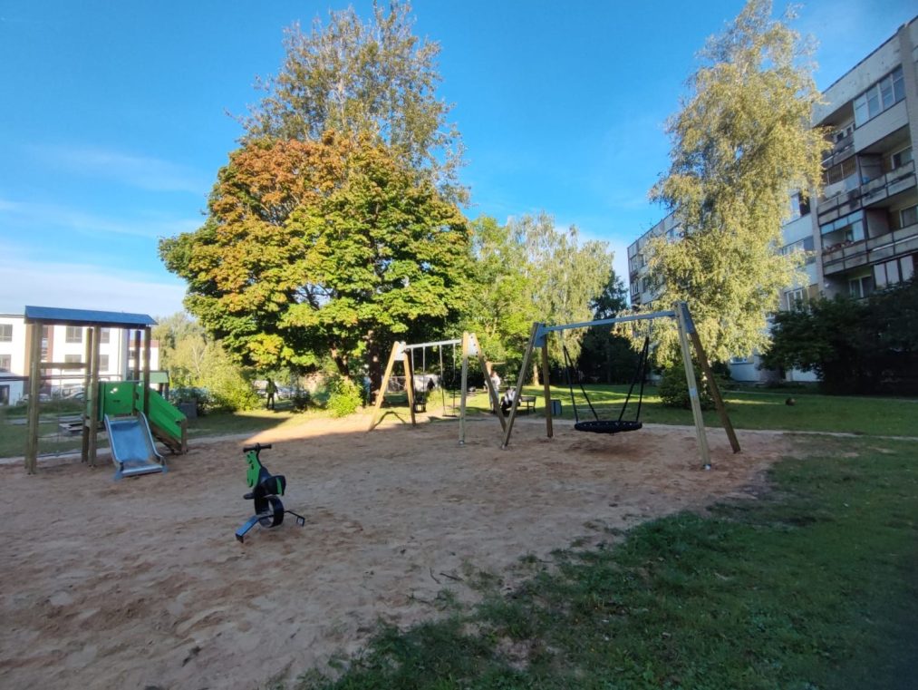 Plašs rotaļu laukums ar šūpolēm, slidkalniņu un līdzsvara velosipēdu smiltīs, fonā koki un mājas.
