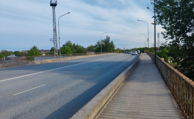 parvads Asfalts pārvads ar gājēju ietvi, metāla margām, apgaismes stabiem, sakaru torni un braucošām automašīnām tālumā.