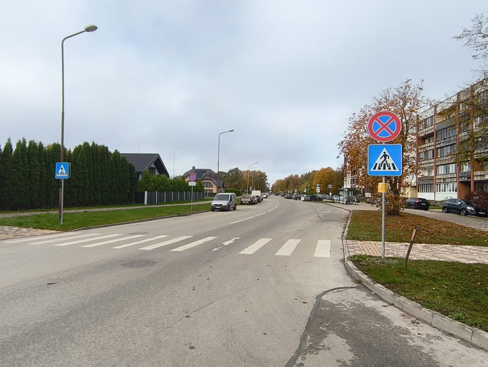 Iela ar gājēju pāreju, ceļa zīmēm un novietotām automašīnām. Labajā pusē daudzdzīvokļu māja.