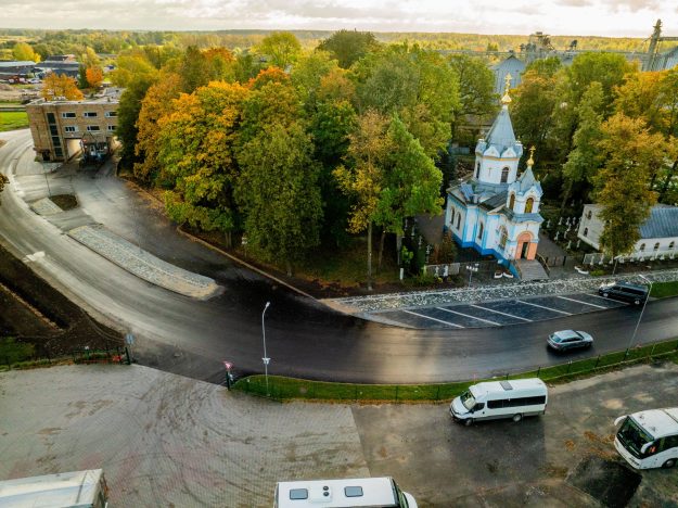 bauskas dzirnavu iela 06 10 2026-08 Skats no augšas uz asfalta ceļu, rudenīgiem kokiem un baltu baznīcu ar zilu jumtu.
