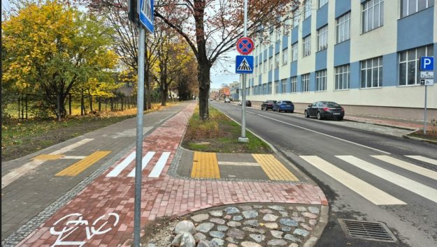 brieza Pilsētas iela rudenī ar gājēju pāreju, veloceliņu, ceļa zīmēm, novietotām automašīnām un daudzstāvu ēku fonā.