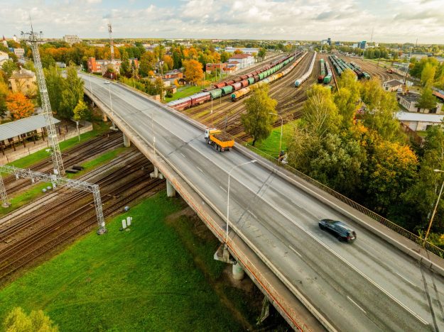 gaisa-tilts-viadukts-dzelscels-lietuvas-soseja-06-10-2026-07-2-scaled Skats no augšas uz pilsētas viaduktu pār dzelzceļa sliežu ceļiem, pa kuru pārvietojas kravas un vieglās automašīnas.