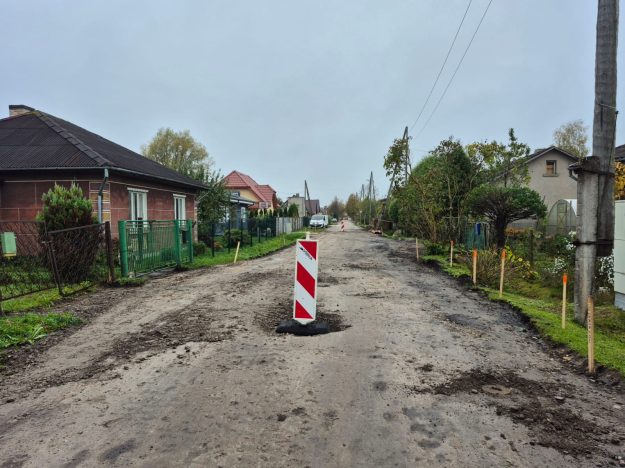 gaismas_iela Zemes ceļš ciematā ar bedrēm un ceļa norobežojošo zīmi priekšplānā. Tālumā redzamas mājas, koki un elektrības stabi.